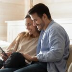 Happy,Man,And,Woman,Using,Phone,Together,,Sitting,On,Couch,