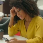 Unhappy,African,American,Woman,In,Cafe,Holding,Head,Smartphone,Mobile